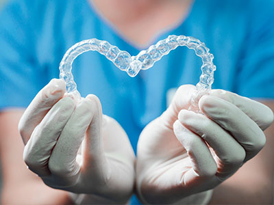 The image shows a pair of hands holding a heart-shaped object made out of clear plastic material, suggesting a theme related to dental care or oral health.