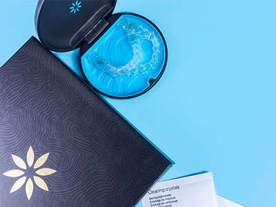 The image shows a close-up view of a cosmetic product, possibly a skincare item, with an open lid revealing its contents, placed on a surface next to a blue cup and a small box with a decorative design.