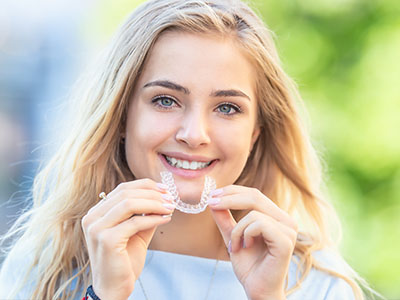 The image features a young woman with blonde hair smiling at the camera while holding up a clear dental retainer.