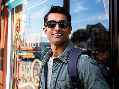 The image shows a man standing outside a storefront, wearing sunglasses and a backpack, with a relaxed posture against a bright background.