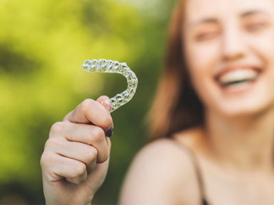 A smiling woman holding up a transparent plastic dental retainer with a smile on her face, set against a blurred outdoor background.